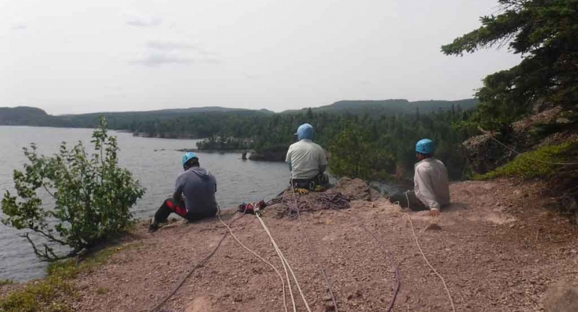 three people sit on a cliff above a body of water and prepare to rock climb on an outward bound expedition 